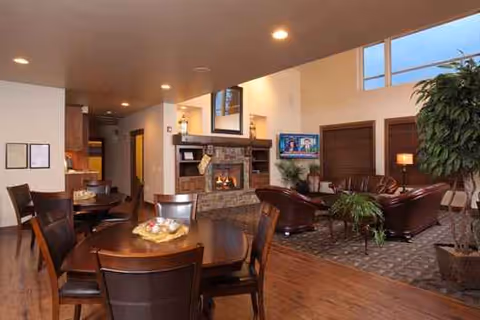 Cozy senior living lounge with fireplace, TV, plants, and wooden tables and chairs.