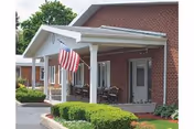 Brick senior living entrance with a covered porch, American flag, and trimmed greenery.