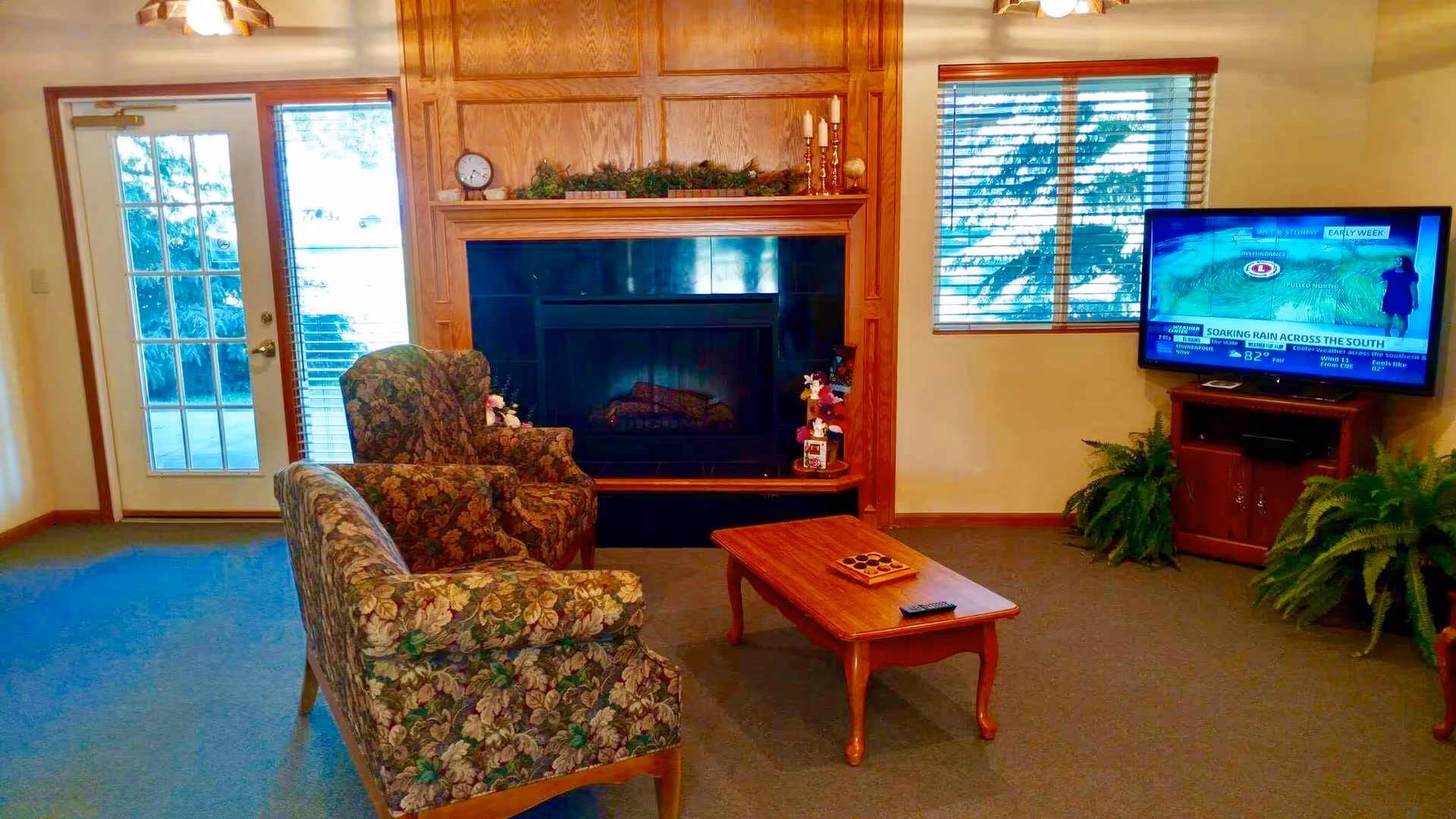 A cozy living room with floral patterned armchairs and a matching loveseat arranged around a wooden coffee table. There is a fireplace with a wooden mantel decorated with greenery and candles. A flat-screen TV is mounted on a wooden stand to the right, with two green potted plants on either side. A door with glass panes and a window with blinds allow natural light into the room.