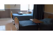 A shared patient room with two hospital-style beds covered in blue bedspreads, bedside tables, tray tables, and windows.