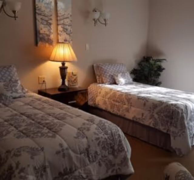 A bedroom with two twin beds covered in matching floral patterned bedding. A nightstand with a lit table lamp is positioned between the beds. Above the nightstand, there are two wall-mounted light fixtures and two decorative wall art pieces. A potted plant is placed in the corner of the room.