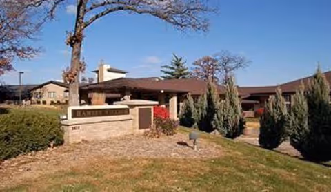 Exterior view of Ramsey Village Continuing Care facility with a stone sign in front, surrounded by grass, shrubs, and trees under a clear blue sky.