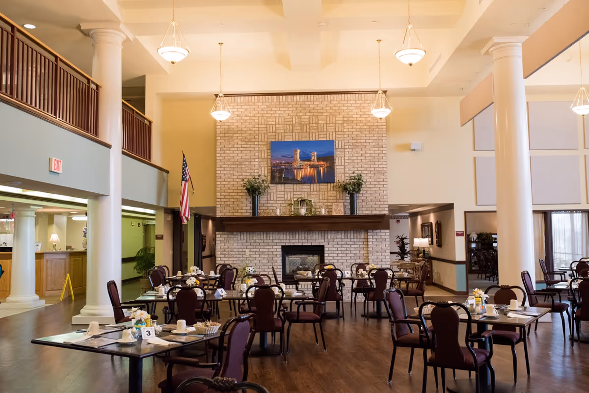 Large communal dining room with multiple set tables and chairs facing a brick fireplace under high ceilings and hanging lights.