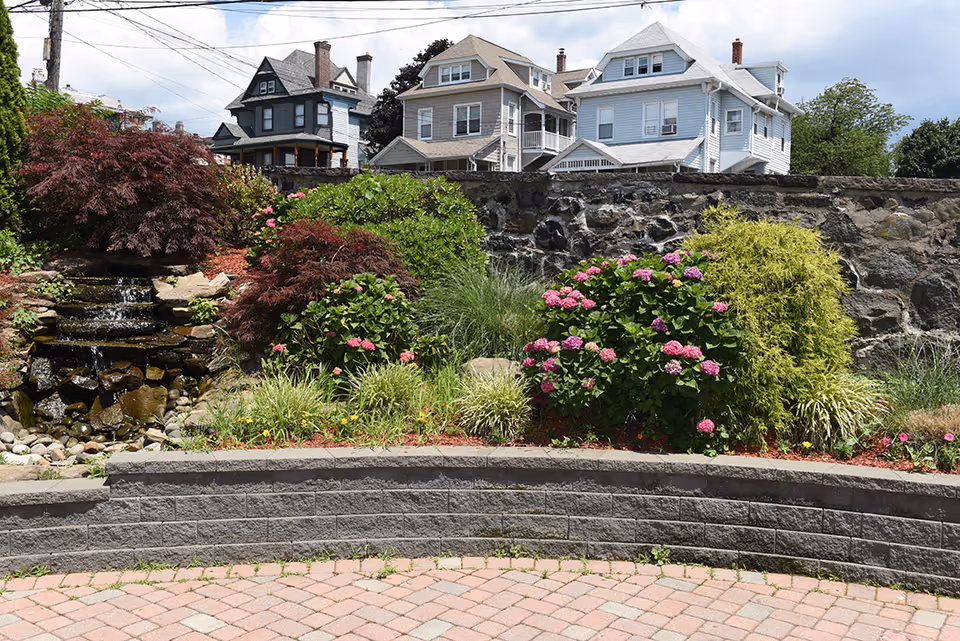 A landscaped outdoor garden area with a small cascading waterfall over rocks, surrounded by various green shrubs and flowering plants including pink and purple hydrangeas. In the background, there is a stone wall and several residential houses under a partly cloudy sky.