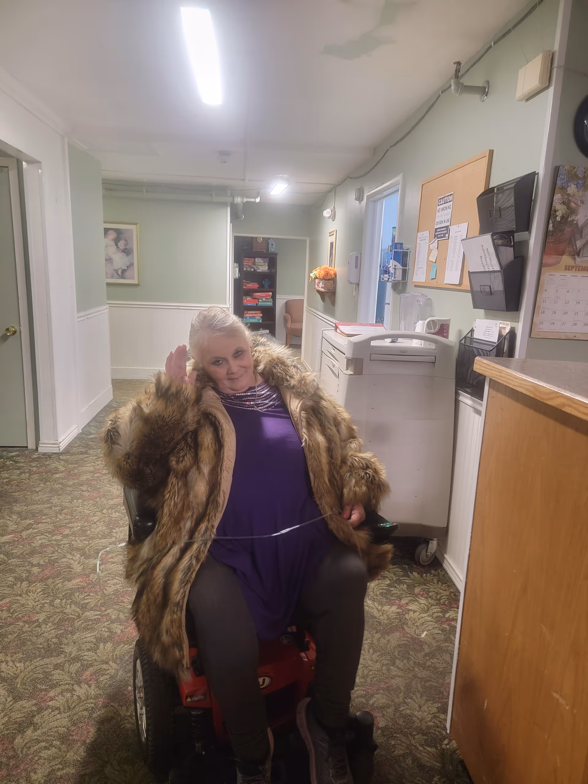 An elderly woman wearing a fur coat and purple dress is sitting on a red mobility scooter in a hallway of an assisted living facility. The hallway has light green walls, floral carpet, and fluorescent ceiling lights. There is a bulletin board, calendar, and some office equipment on the right side of the hallway.