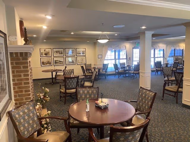 A spacious dining room in a senior living facility with multiple round wooden tables and cushioned chairs arranged neatly. The room has carpeted floors, beige walls adorned with framed pictures, and large windows with sheer curtains allowing natural light to enter. A small flower arrangement and a bottle of hand sanitizer are placed on the nearest table.