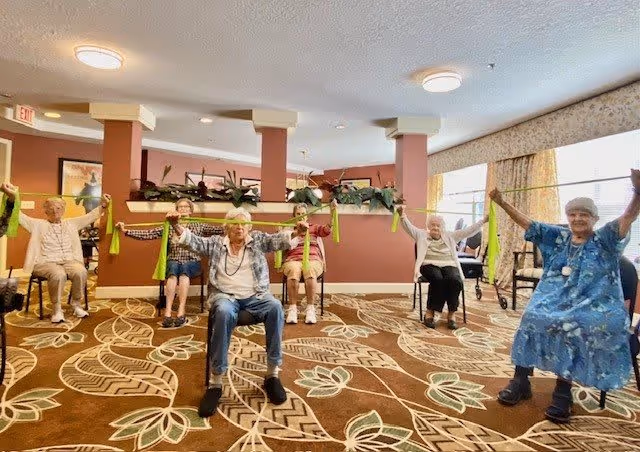A group of elderly individuals seated in a semi-circle in a common room, each holding up a green exercise band as part of a group activity. The room has a patterned carpet, beige walls with a reddish-brown accent, and large windows with curtains allowing natural light in.