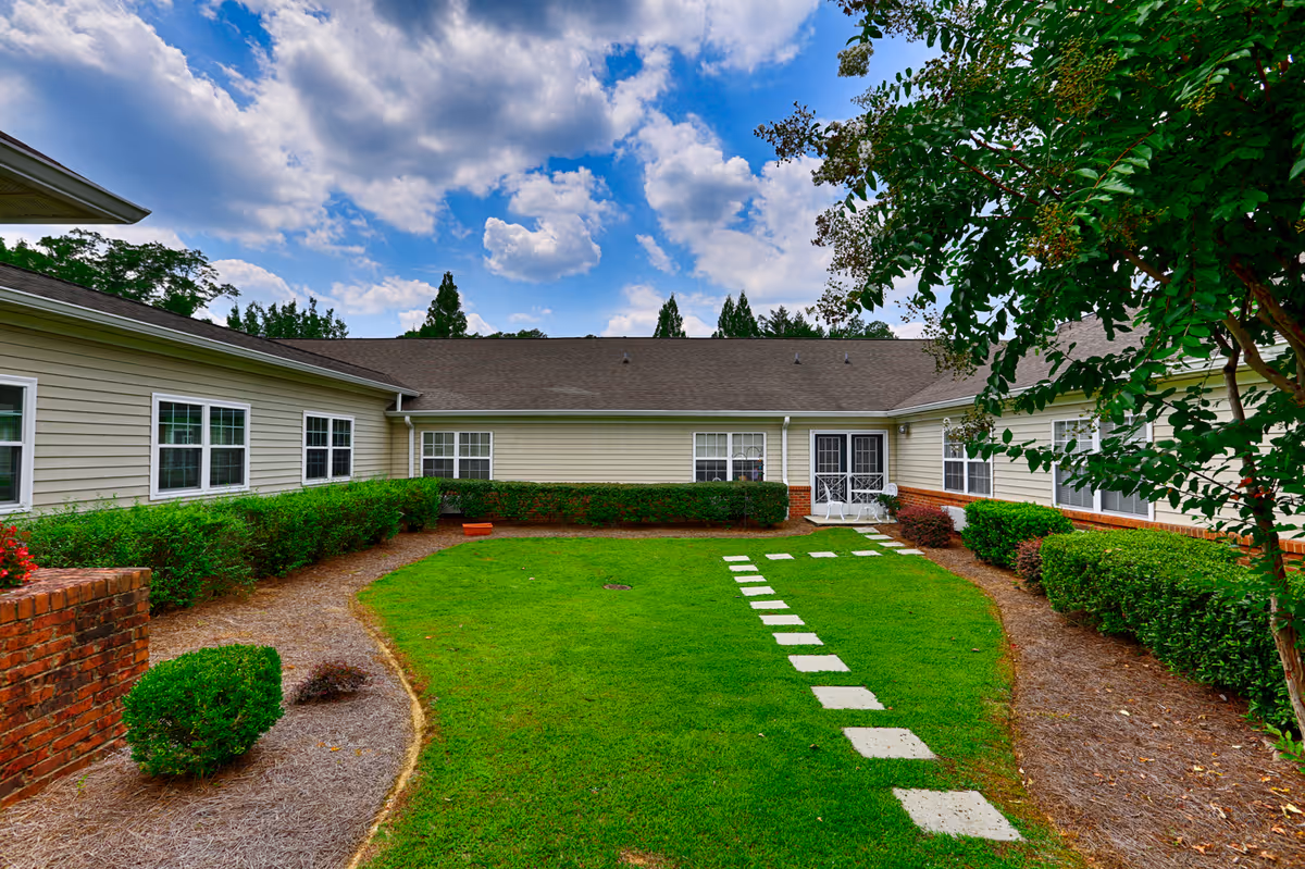 A well-maintained outdoor courtyard area with a green lawn and a stone pathway leading to a white double door entrance. The courtyard is surrounded by beige buildings with white-framed windows, trimmed bushes, and a tree on the right side. The sky is partly cloudy with blue patches visible.
