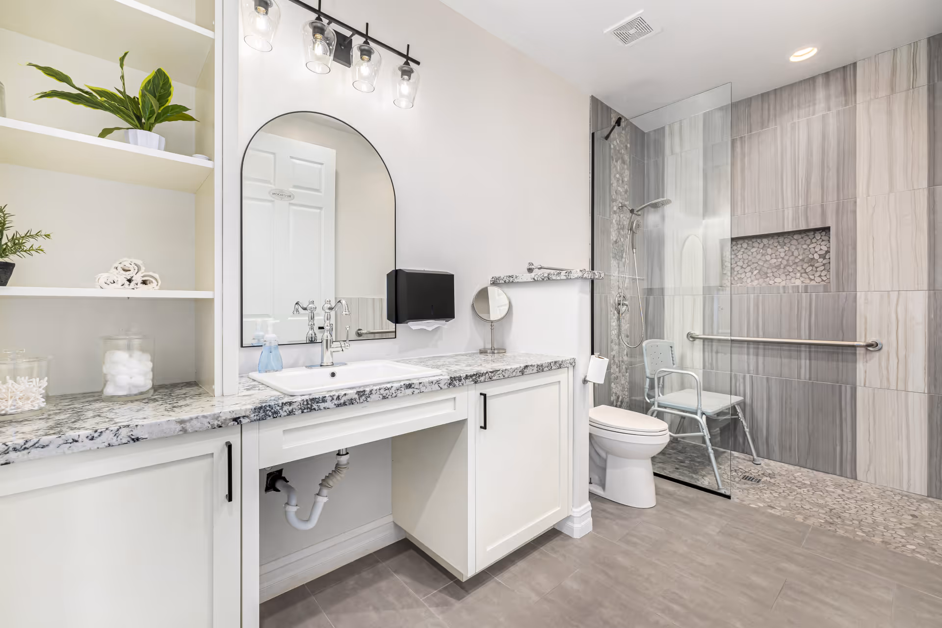A modern bathroom with a granite countertop, white cabinetry, a large arched mirror, and a sink with a soap dispenser. There are shelves with plants and cotton balls on the left. The bathroom features a walk-in shower with a glass partition, a shower chair, grab bars, and pebble tile accents. The floor is tiled in gray, and the walls are light-colored.