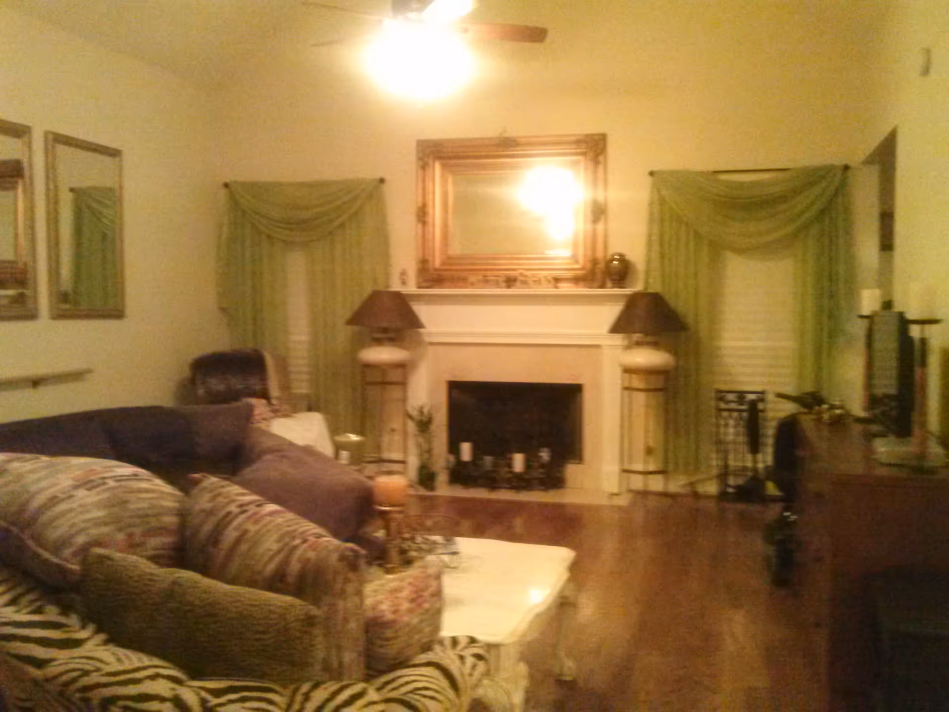 A cozy living room with a wooden floor, a fireplace in the center with a large framed mirror above it, two tall lamps on either side of the fireplace, green curtains on the windows, a patterned sofa with multiple cushions, a white coffee table, and a ceiling fan with a light.