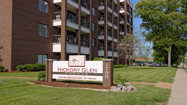 Exterior view of a multi-story brick senior living facility named Hickory Glen with balconies. In front of the building is a large sign displaying the facility's name, address, and phone number. The surrounding area has green grass, trees, and a sidewalk.