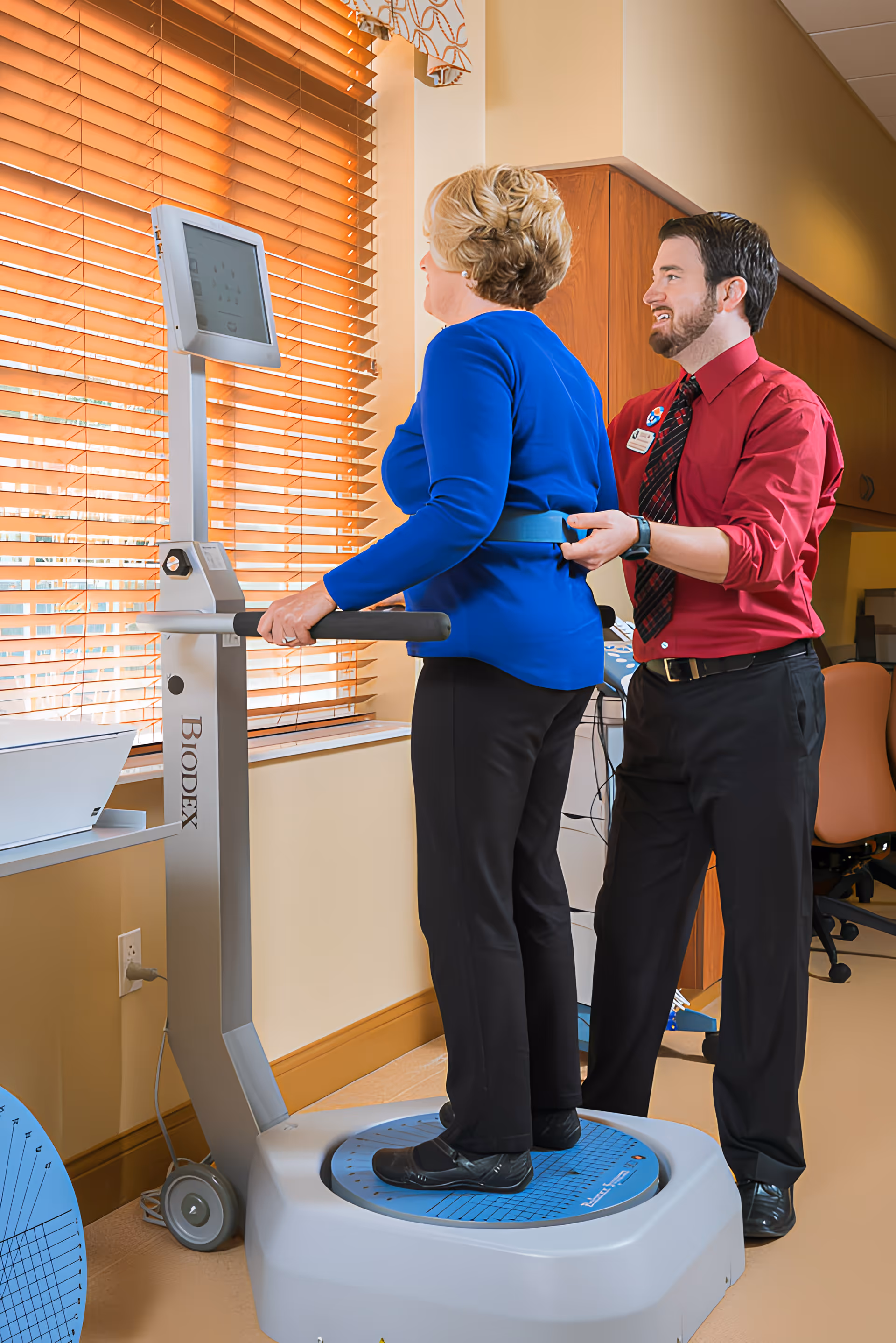 A woman stands on a Biodex balance testing machine holding the handles while a man in a red shirt and black pants assists her by adjusting a belt around her waist. They are in a well-lit room with wooden blinds on the window and office furniture in the background.