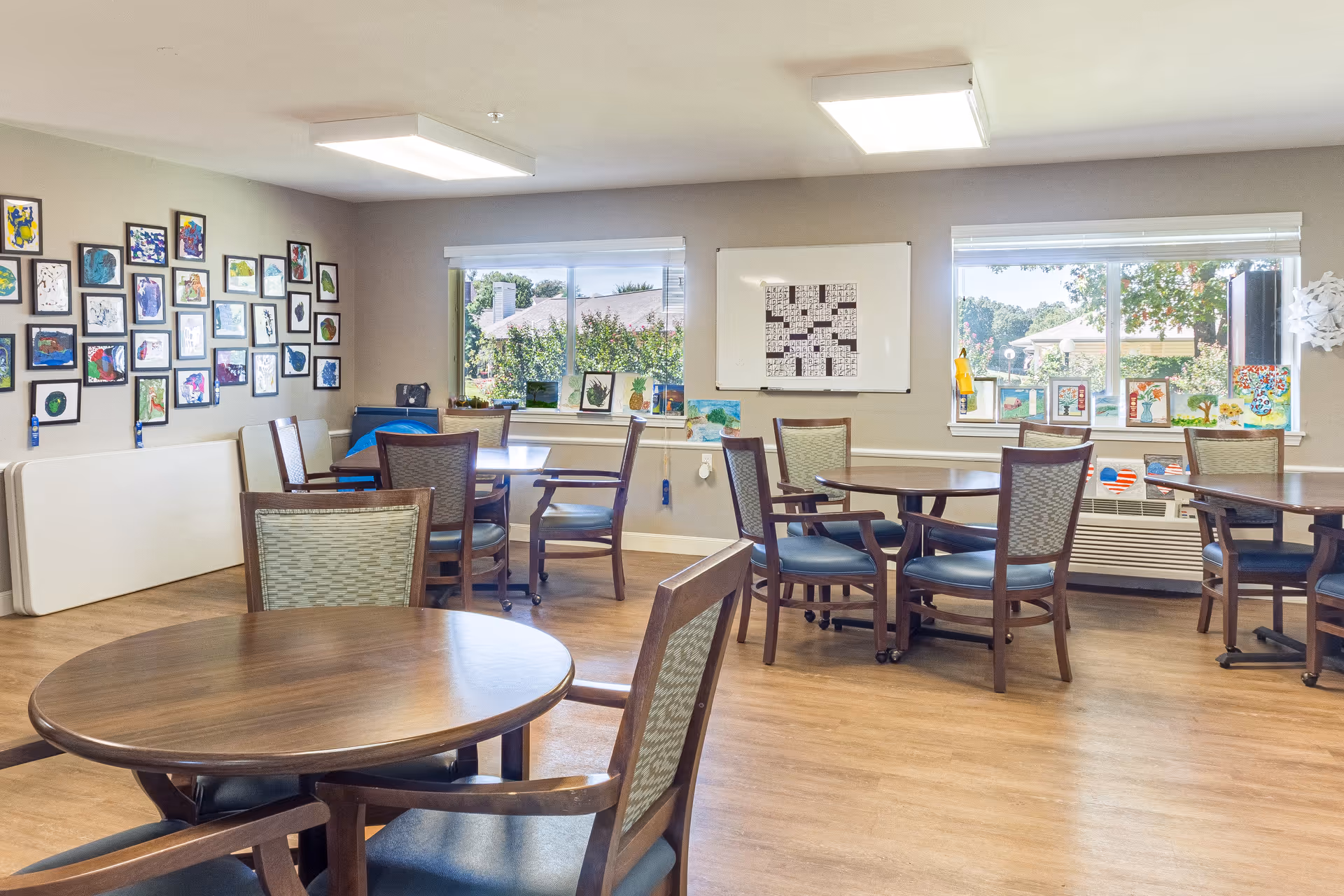 Bright communal dining/activity room with round wooden tables and chairs, framed artwork on the wall, and windows looking outside.