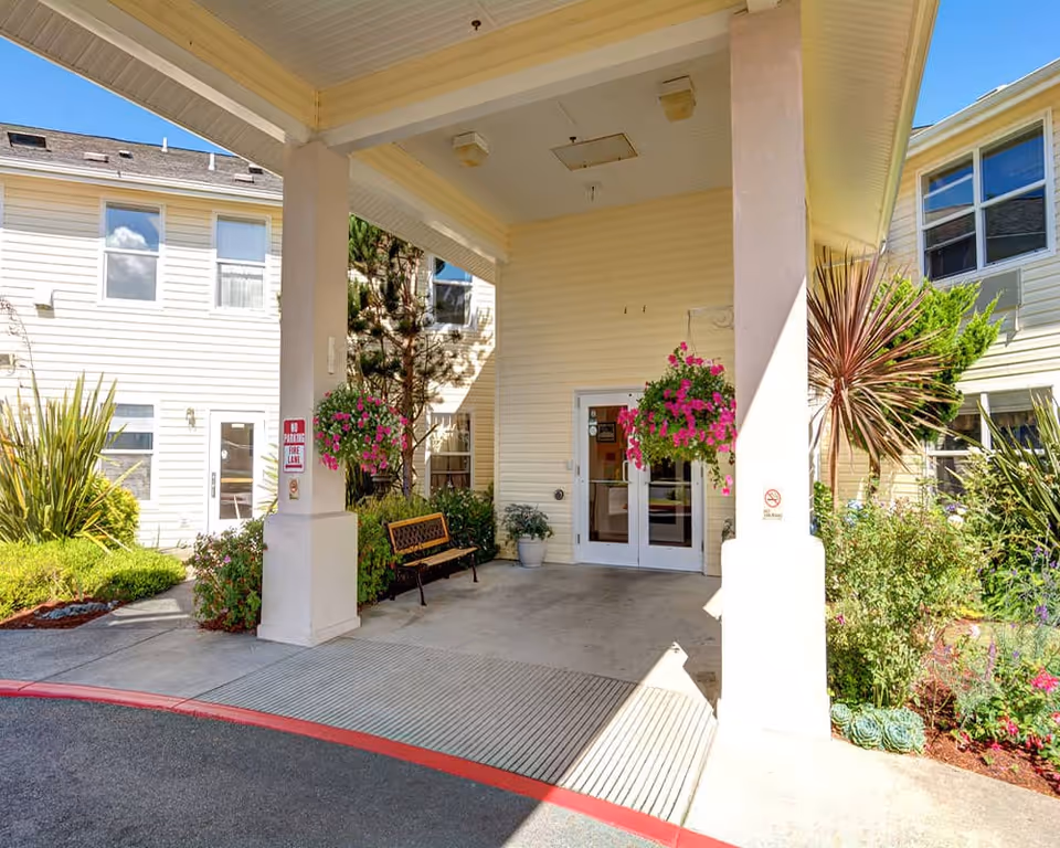 Covered entrance area of a senior living facility with two white pillars, hanging flower baskets, a bench, and glass double doors. The building exterior is light yellow with multiple windows, surrounded by green plants and flowers.