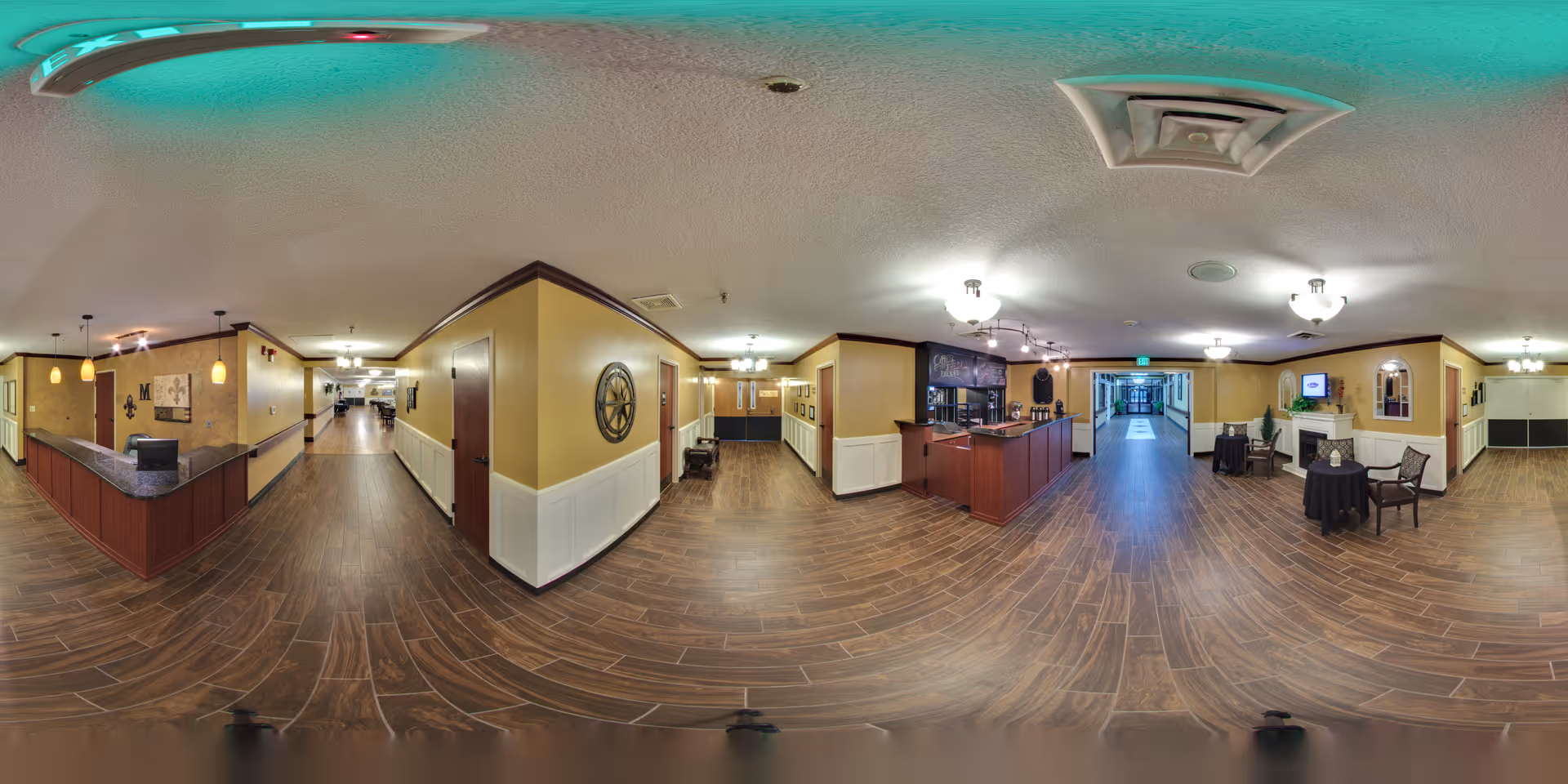 Panoramic view of a senior living facility interior hallway with wood flooring, beige walls, and white wainscoting. The area includes a reception desk on the left, a small seating area with chairs and a table on the right, and a hallway leading to other rooms and areas. Ceiling lights and decorative elements are visible throughout the space.