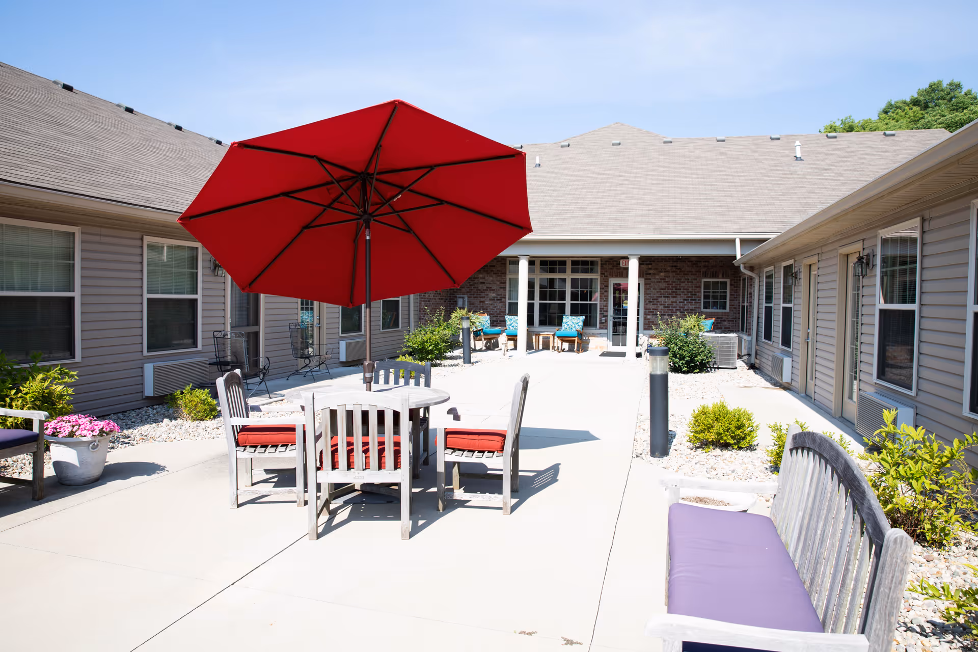 Outdoor courtyard area at Brownsburg Meadows Assisted Living with a round table and four chairs under a large red umbrella. There are additional benches with cushions and some potted plants around the concrete patio. The building surrounds the courtyard with windows and doors visible.