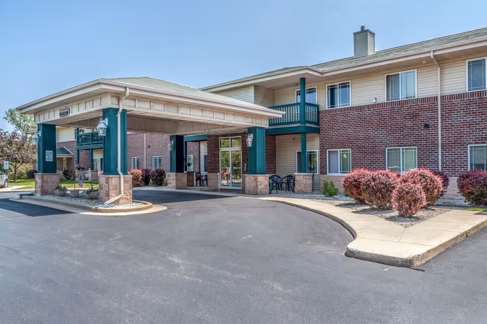 Exterior view of Prairie Meadows facility showing a two-story brick and siding building with a covered entrance driveway. There are windows on both floors, a balcony, outdoor chairs, and landscaped bushes along the sidewalk.