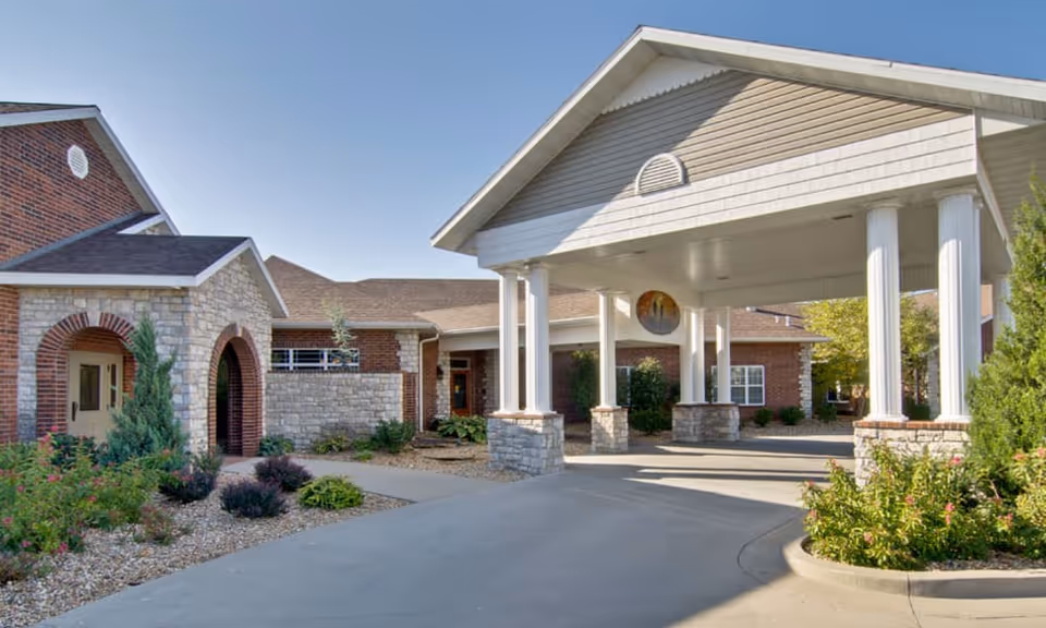 Front entrance of a brick-and-stone senior living building with a covered porte-cochere supported by white columns and a landscaped driveway.