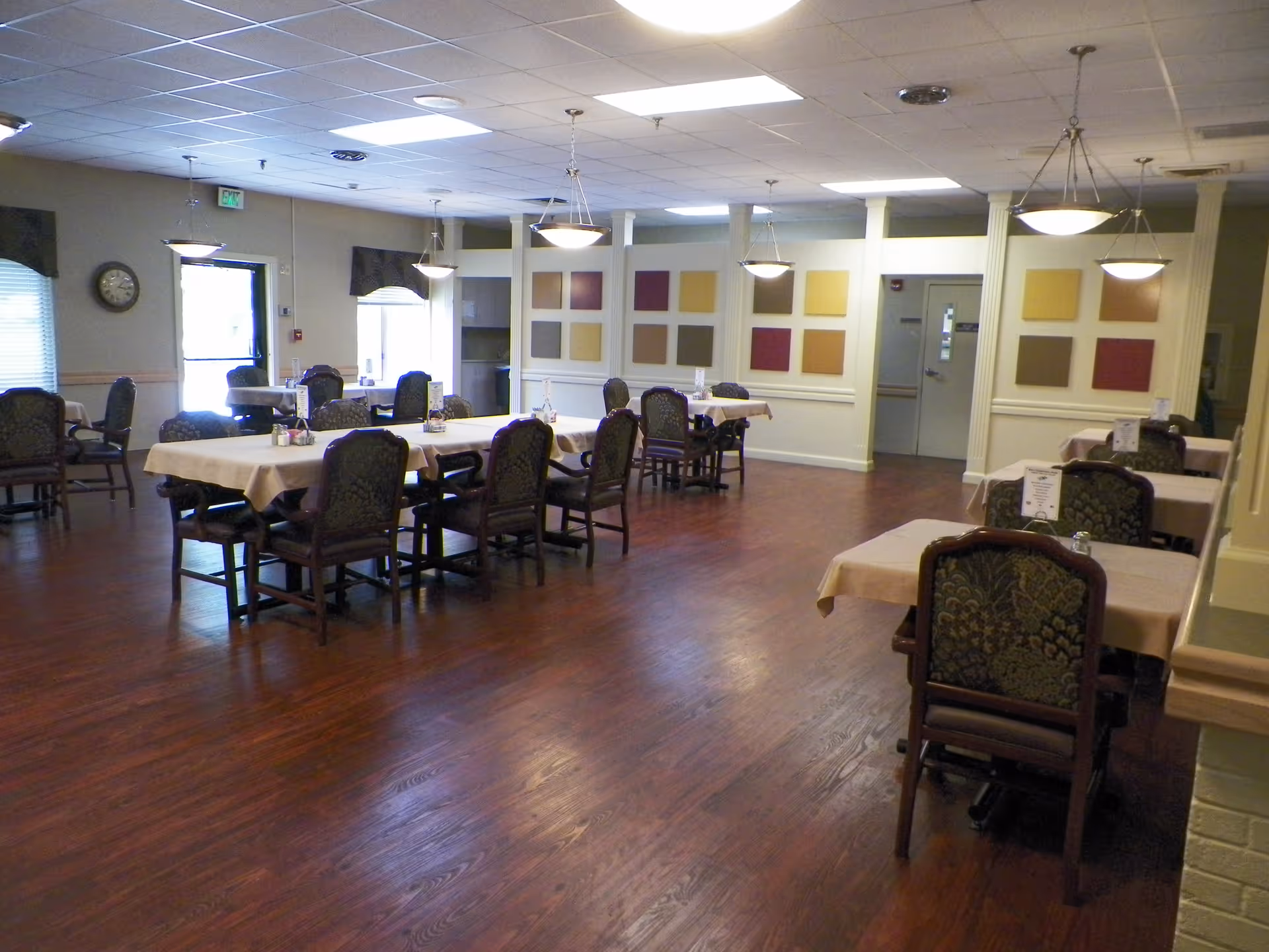 A dining room with several tables covered with beige tablecloths and surrounded by upholstered chairs. The room has wooden flooring, hanging pendant lights, and decorative square wall panels in various colors. There is a clock on the wall near a door with a window, and windows with blinds letting in natural light.