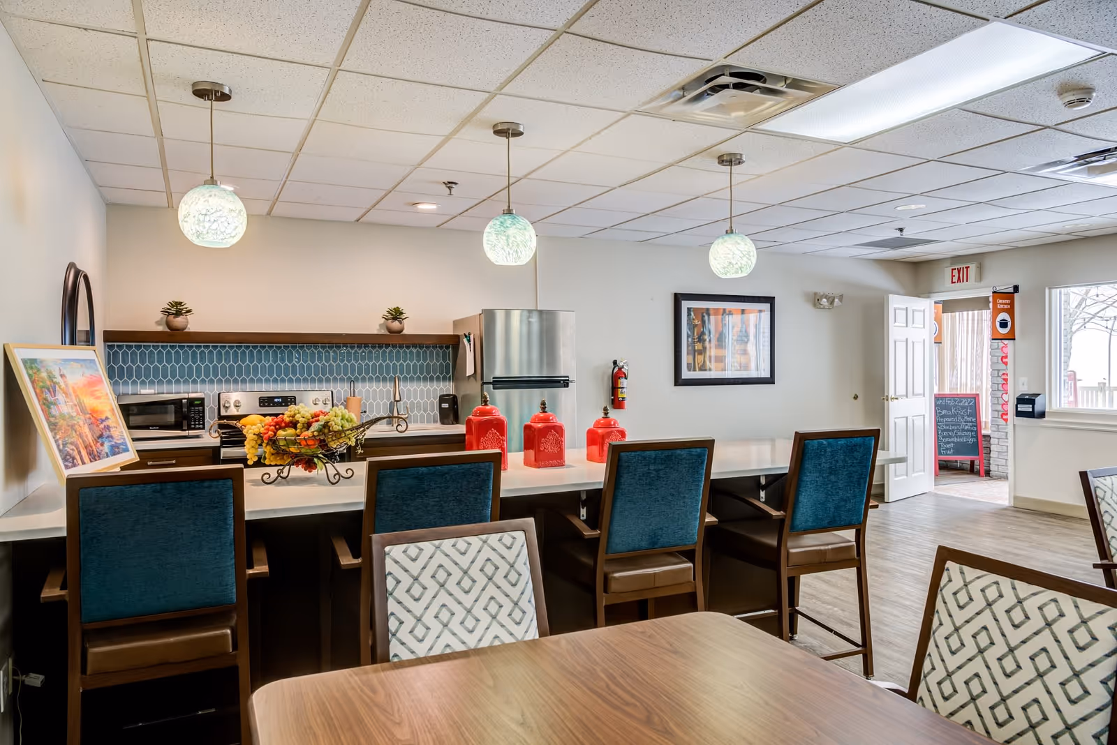 Interior view of a senior residence kitchen and dining area featuring a long counter with four blue cushioned chairs, three hanging pendant lights, a stainless steel refrigerator, stove, microwave, and a decorative fruit basket on the counter. There are red canisters on the counter, a framed picture on the wall, and a table with patterned chairs in the foreground. An exit door and a chalkboard menu are visible in the background.
