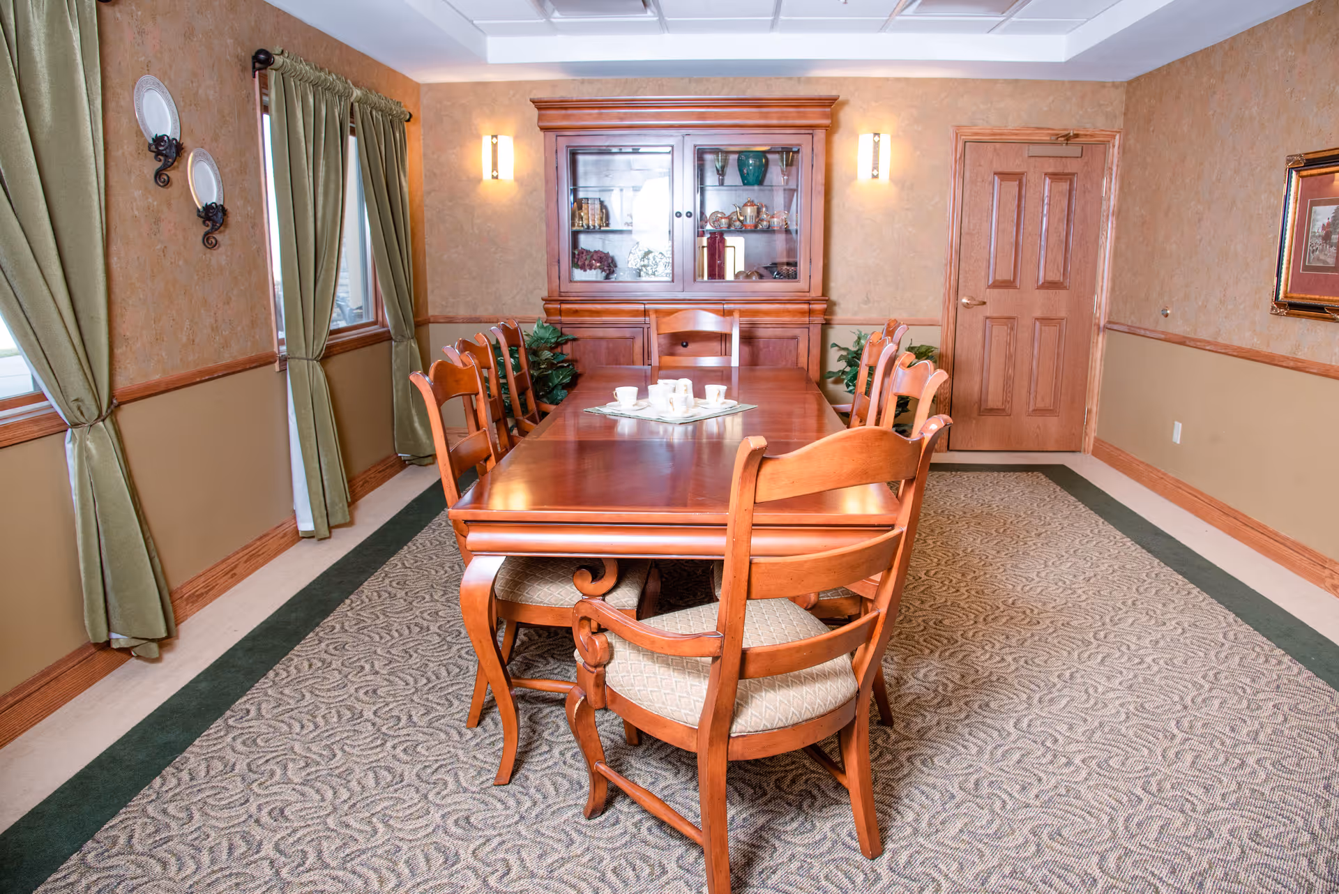 A formal dining room with a long wooden table surrounded by eight wooden chairs with cushioned seats. The room features beige walls with a wooden chair rail, green curtains on two windows, a patterned carpet, a wooden cabinet with glass doors displaying decorative items, and wall sconces providing lighting.