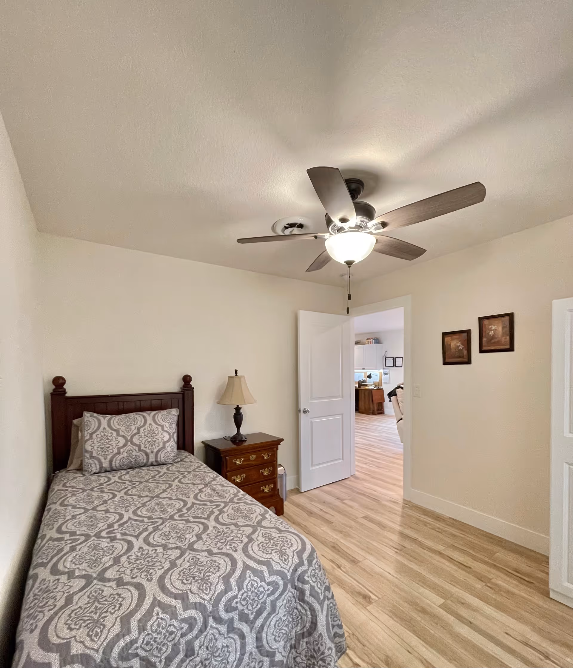 A small bedroom with a single bed covered in a gray and white patterned bedspread. Next to the bed is a wooden nightstand with a lamp. The room has light-colored walls and wood flooring. A ceiling fan with a light is mounted on the ceiling. The door to the room is open, showing a glimpse of another room with similar flooring and furniture.