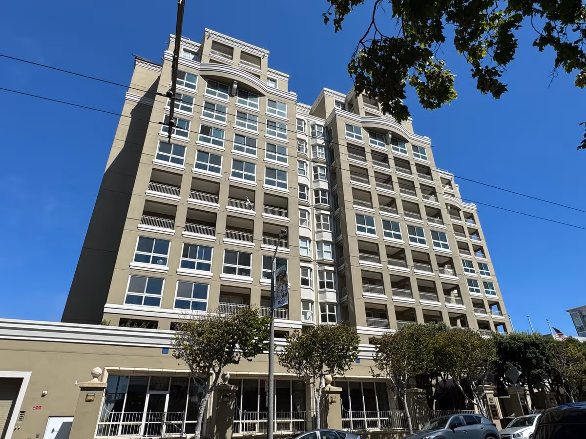 Front exterior of a multi-story residential building with balconies, trees, and parked cars under a clear blue sky.