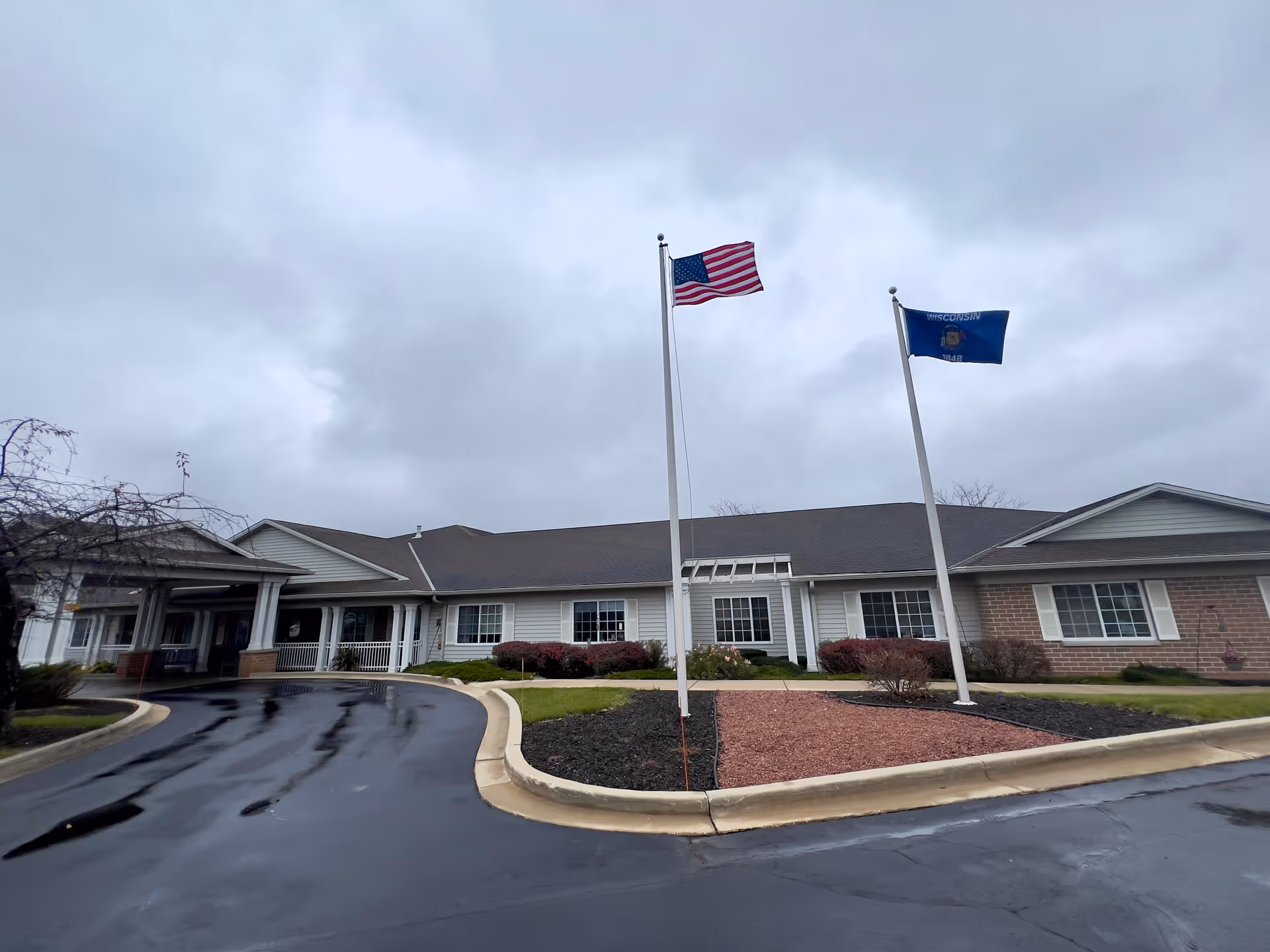 Exterior view of Brenwood Park Assisted Living facility on a cloudy day, showing a single-story building with white siding and brick accents, two flagpoles with the American flag and Wisconsin state flag, and a curved driveway in front.