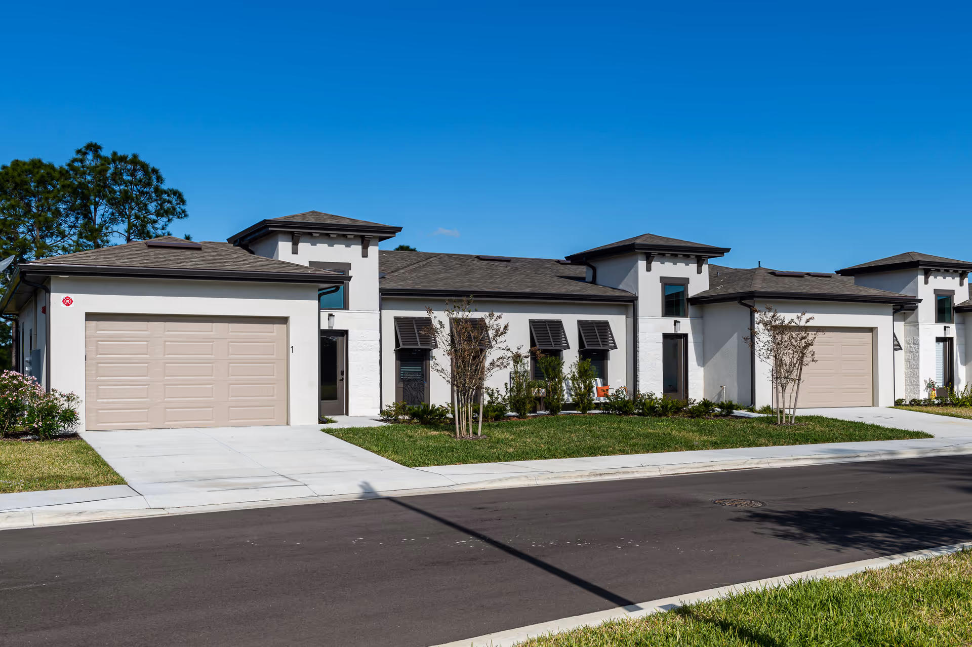 Exterior view of a modern single-story senior living facility building with multiple attached units, each featuring a garage, windows with awnings, and small landscaped areas with grass and young trees under a clear blue sky.
