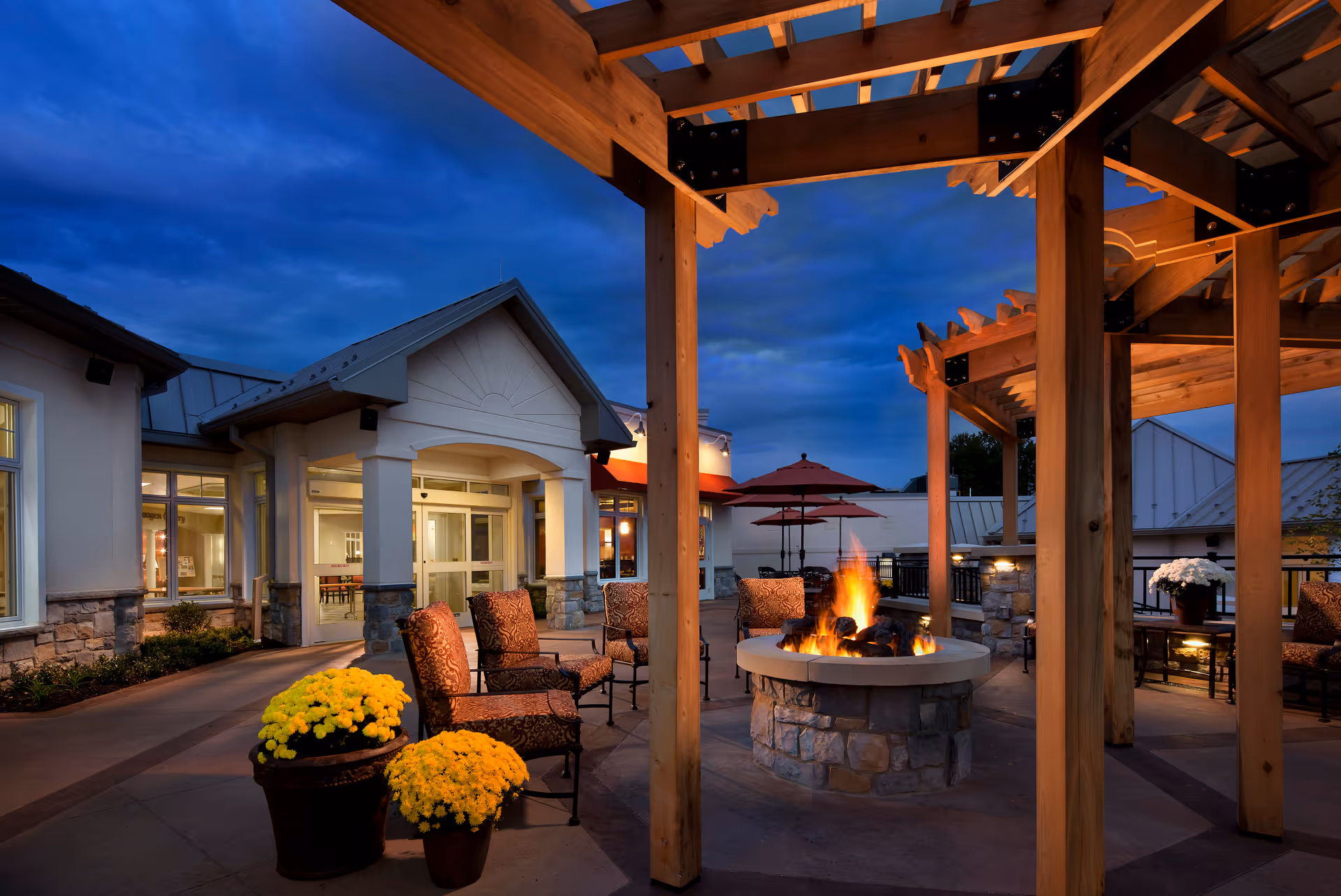 Outdoor patio area at dusk with a stone fire pit surrounded by cushioned chairs under wooden pergolas. Yellow flowers in pots are placed near the seating area, and the building entrance with large windows and a peaked roof is visible in the background.