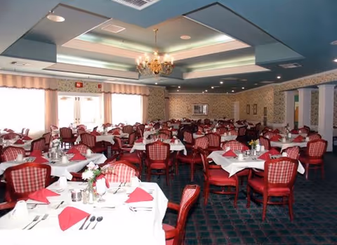A spacious dining room with multiple tables covered in white tablecloths and red napkins. Each table is set with silverware, glasses, and small floral centerpieces. The room features patterned wallpaper, a chandelier hanging from a recessed ceiling, and large windows letting in natural light.