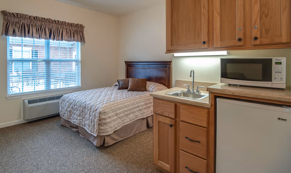 A senior living facility room featuring a bed with a wooden headboard and two brown pillows, a window with blinds and a brown valance, a small kitchenette area with a sink, wooden cabinets, a microwave, and a mini refrigerator.