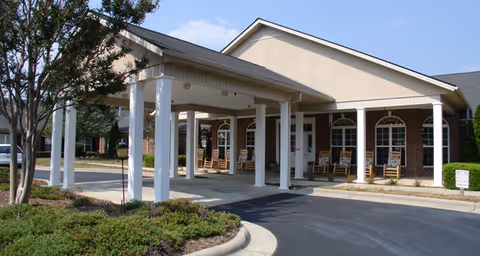 Entrance of a senior living facility with a covered driveway supported by white columns. There are rocking chairs on the porch area in front of the building, which has brick and beige siding. A tree and some shrubs are visible in the landscaped area near the driveway.
