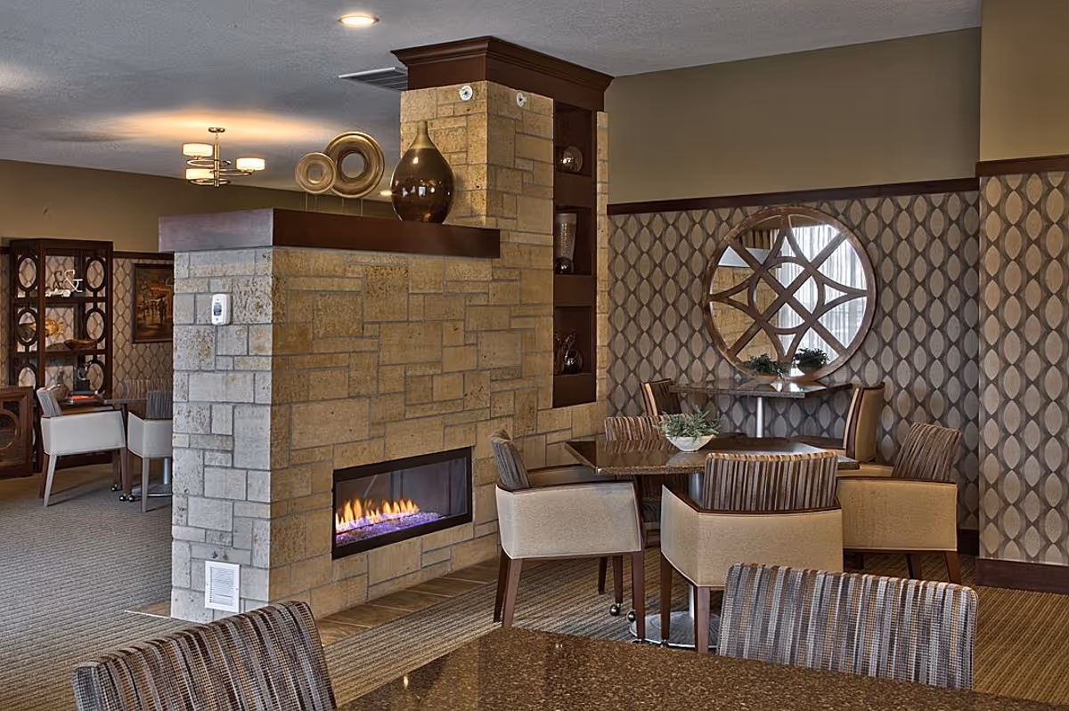 A cozy interior common area with a stone fireplace featuring a modern gas fire. Surrounding the fireplace are several tables with cushioned chairs, decorative vases on the mantel, and a round mirror with a geometric wooden frame on the patterned wallpapered wall.