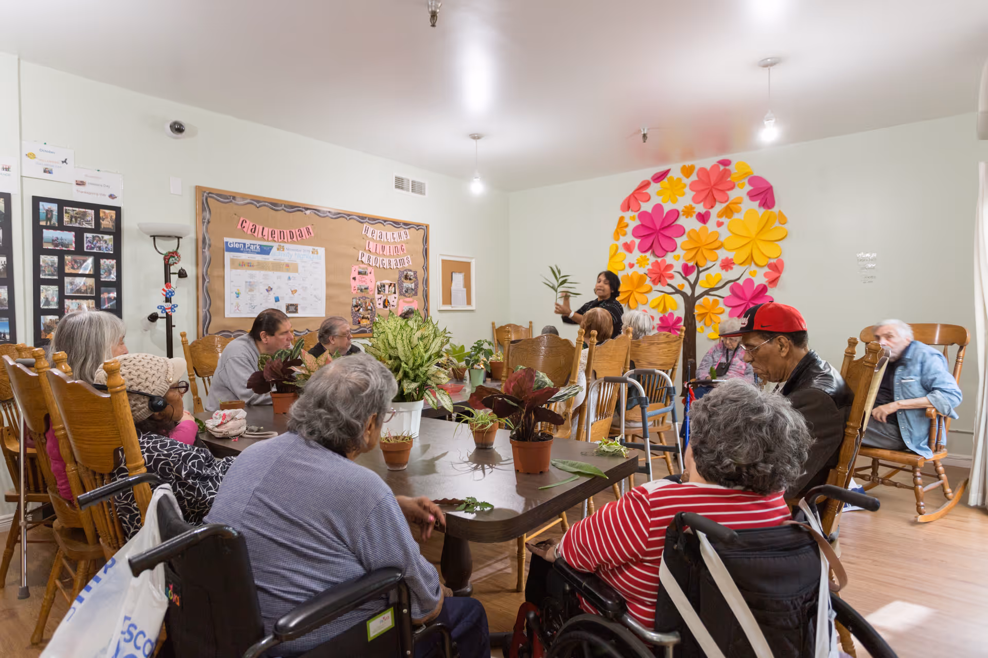 A group of elderly people seated around a large table in a community room, engaging in a plant-related activity led by a standing instructor. The room has wooden chairs, potted plants on the table, a colorful flower mural on the wall, and a bulletin board with calendars and health program information.