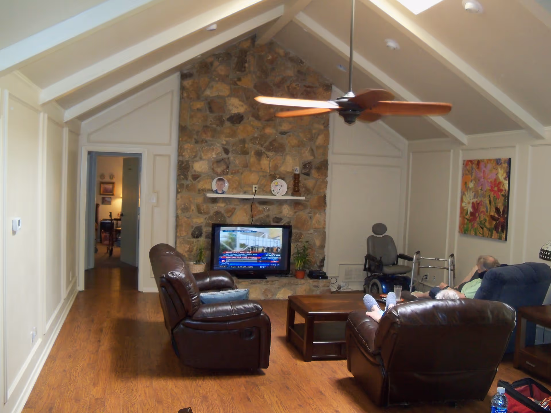 Vaulted living room with a stone fireplace and TV, leather recliners, ceiling fan, coffee table, and mobility aids.
