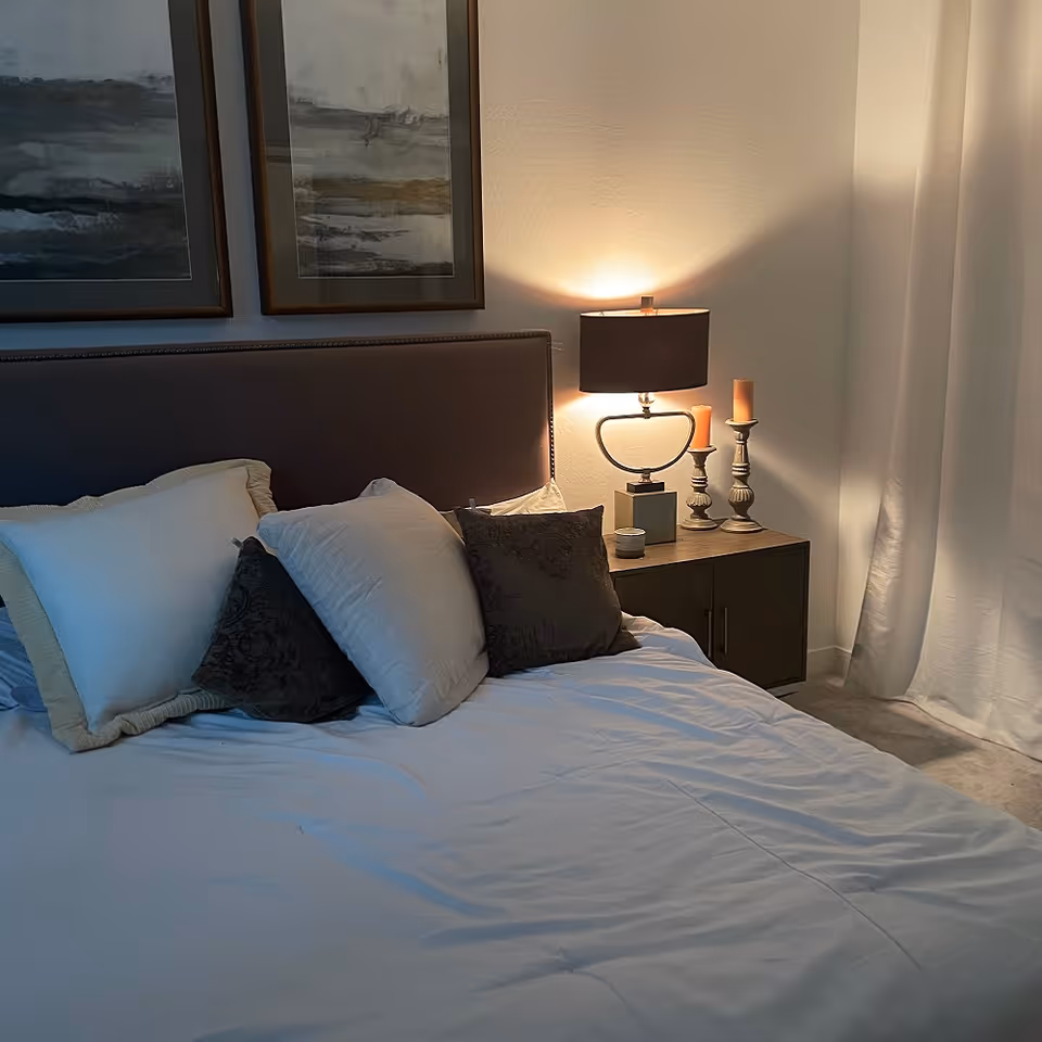 A cozy bedroom with a neatly made bed featuring multiple pillows in white and dark colors. There is a dark upholstered headboard, two framed abstract paintings on the wall above the bed, and a wooden nightstand with a modern lamp and two decorative candles. White curtains hang to the right side of the nightstand.