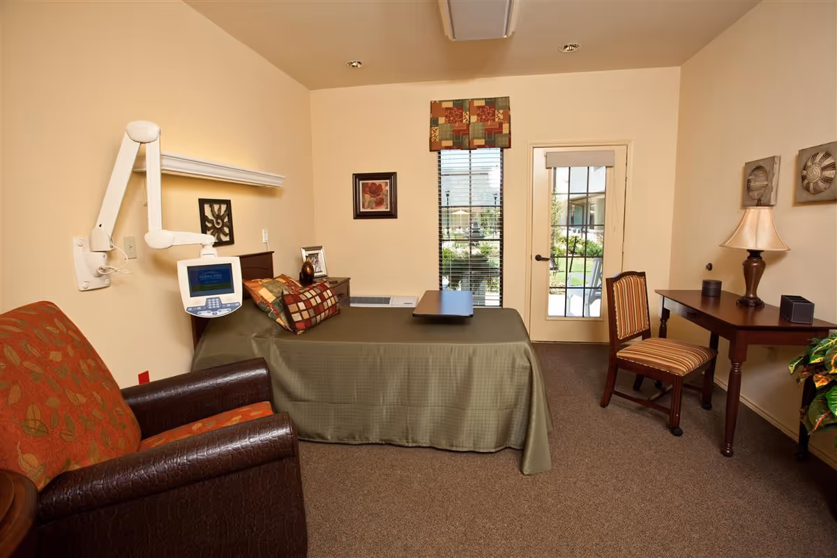 A cozy bedroom in a senior living facility with a single bed covered in an olive green bedspread and decorative pillows. There is a medical monitor mounted on the wall next to the bed. A comfortable armchair with a patterned orange cushion is positioned near the bed. A wooden desk with a lamp and a striped chair is placed against the wall. A window with blinds and a glass door lead to an outdoor garden area. The room has beige walls and carpeted flooring.