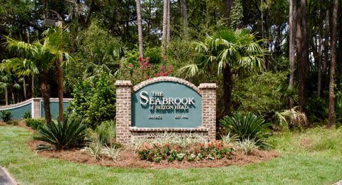 A landscaped entrance sign for The Seabrook of Hilton Head, featuring a brick base and green sign surrounded by palm trees, bushes, and other greenery with a wooded area in the background.