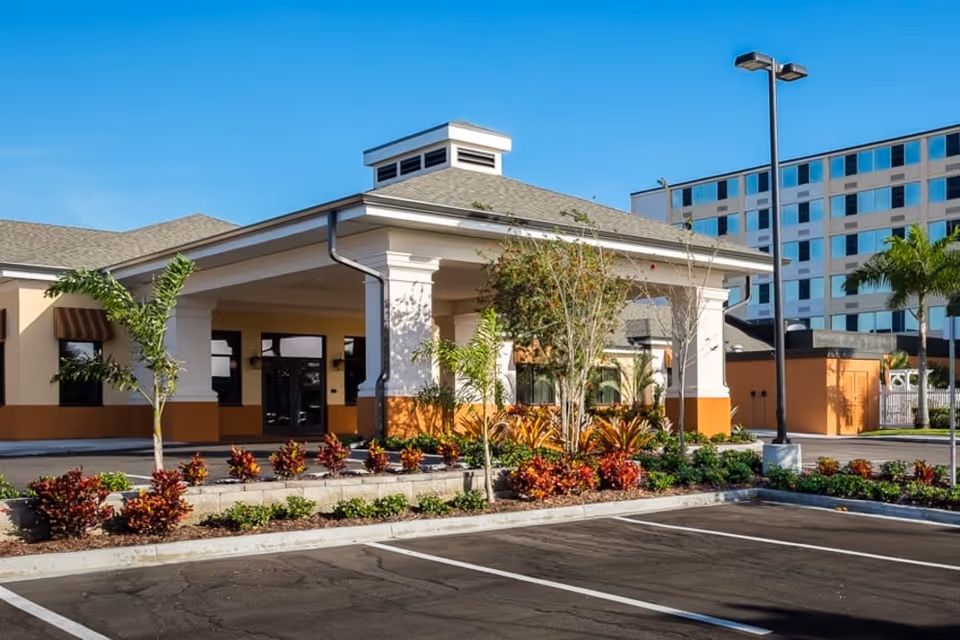 Front entrance and porte-cochere of a senior living facility with landscaped beds and empty parking spaces under a clear blue sky.
