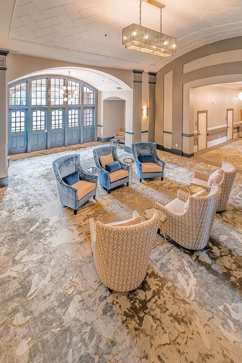 A spacious senior living facility common area with a patterned carpet and a seating arrangement consisting of three blue armchairs and four beige patterned armchairs around small round tables. The room features large arched windows with blue frames, a modern rectangular chandelier hanging from a white ceiling, and a hallway with doors visible in the background.