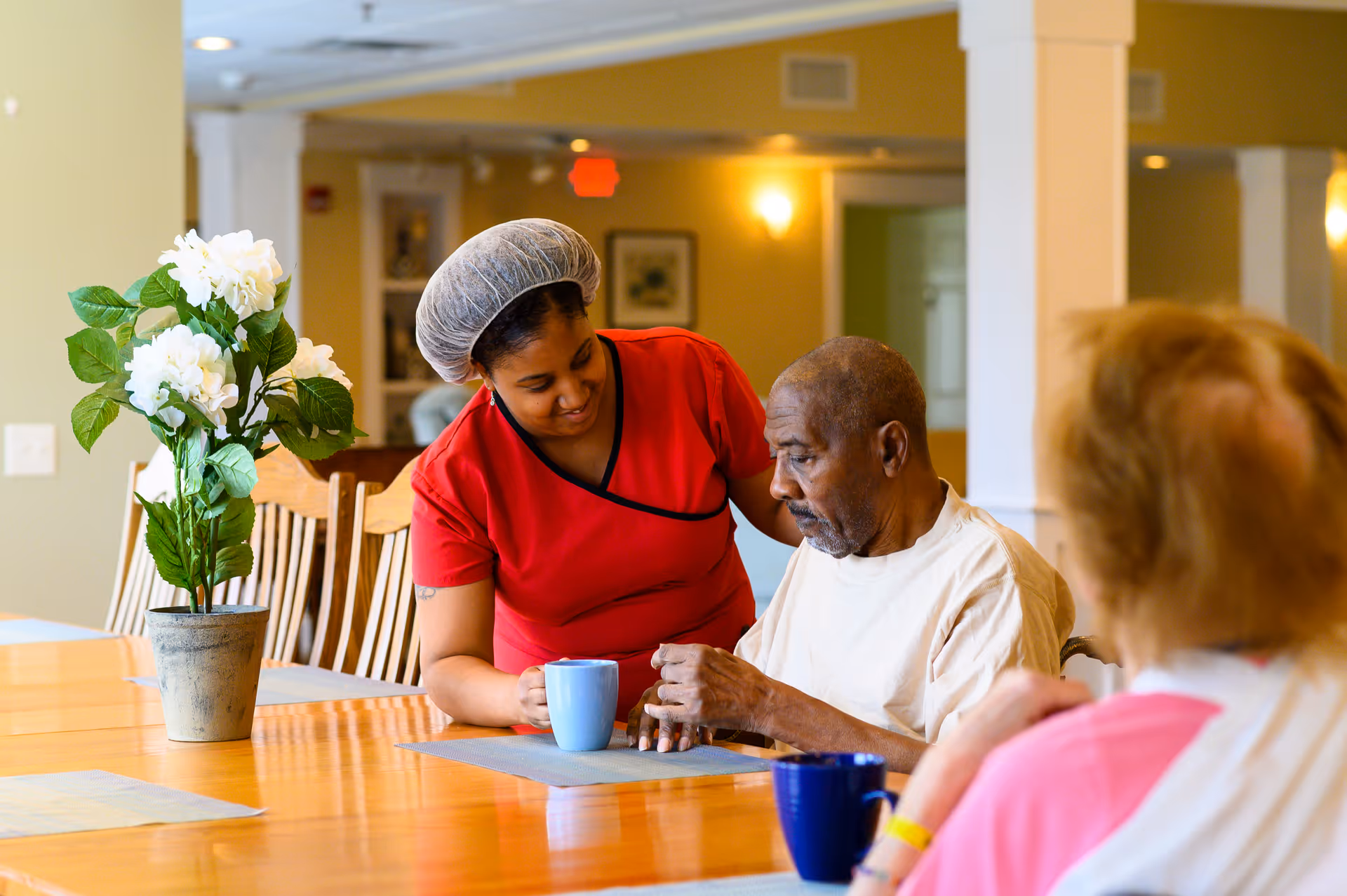 A caregiver in a red uniform and hairnet is assisting an elderly man seated at a wooden table in a well-lit room. The man is holding a blue mug, and another elderly person with reddish hair and a pink shirt is seated nearby. A potted plant with white flowers is on the table.
