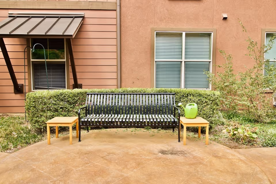 Black metal outdoor bench between two small wooden tables (one with a green watering can) in front of hedges and a building with windows.