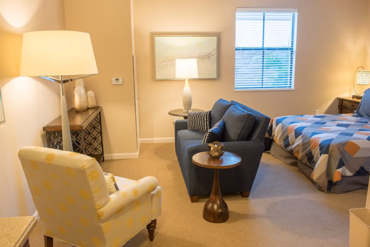 A cozy bedroom with a bed covered in a geometric patterned quilt, a blue loveseat with decorative pillows, a yellow armchair with a circular pattern, two side tables with lamps, a framed artwork on the wall, and a window with blinds letting in natural light.