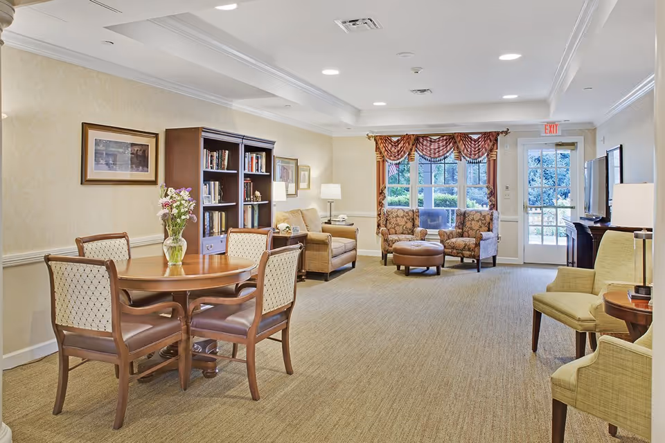 A bright and spacious living room area in a senior living facility with a round wooden table and four chairs, a bookshelf filled with books, comfortable armchairs, a large window with decorative curtains, and a door leading outside. The room is well-lit with ceiling lights and lamps.