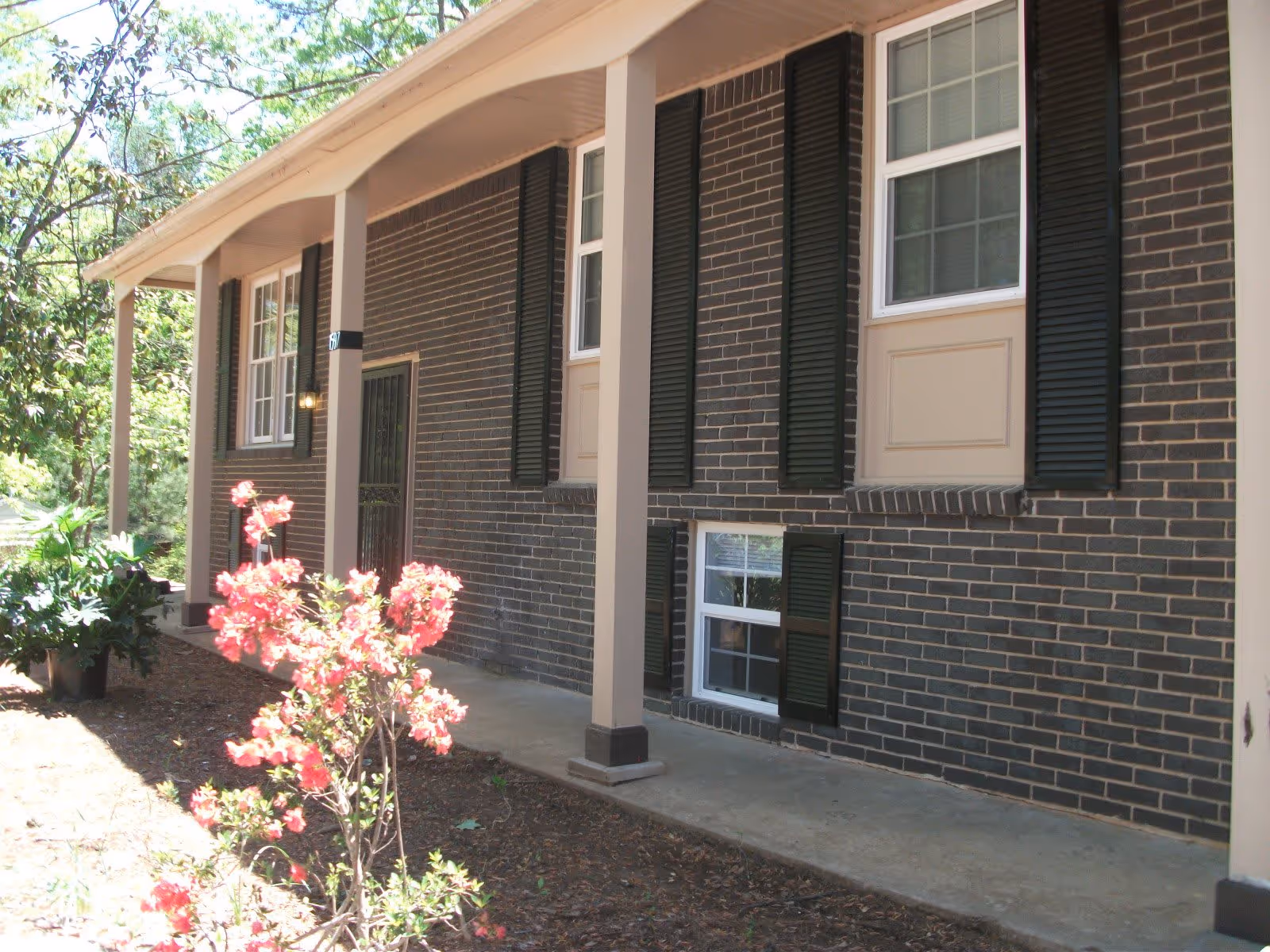 Exterior view of a brick building with black shutters on the windows and a covered porch supported by beige columns. There are pink flowering plants and greenery in the garden area beside the porch.