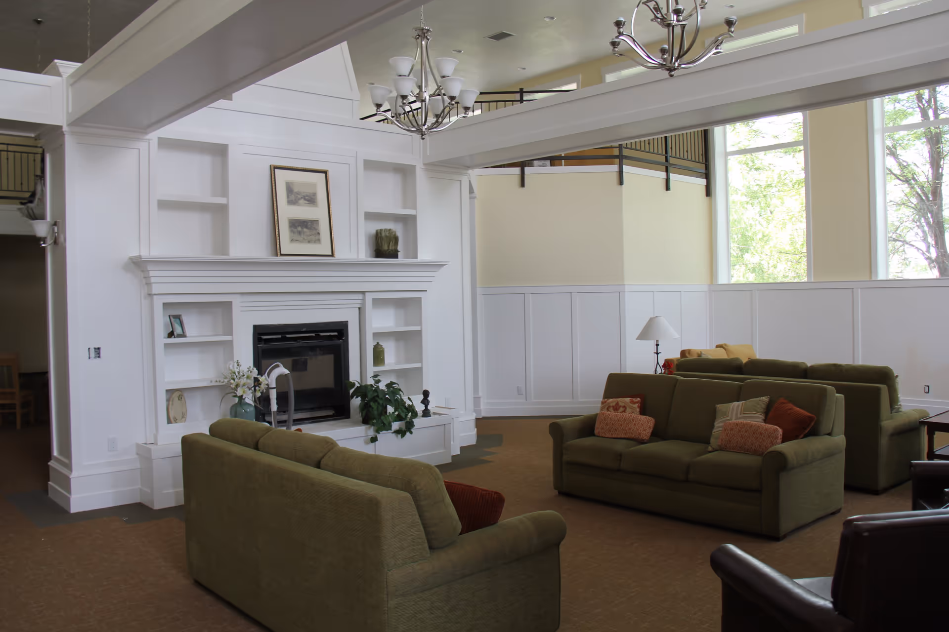 Spacious common lounge with green sofas facing a white built-in fireplace and large windows.