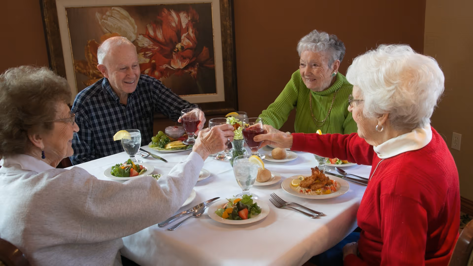Four elderly people sitting around a dining table in a retirement community, raising their glasses in a toast. The table is set with plates of food including salad, bread rolls, and a main dish, along with glasses of water and juice. A floral painting is visible on the wall behind them.