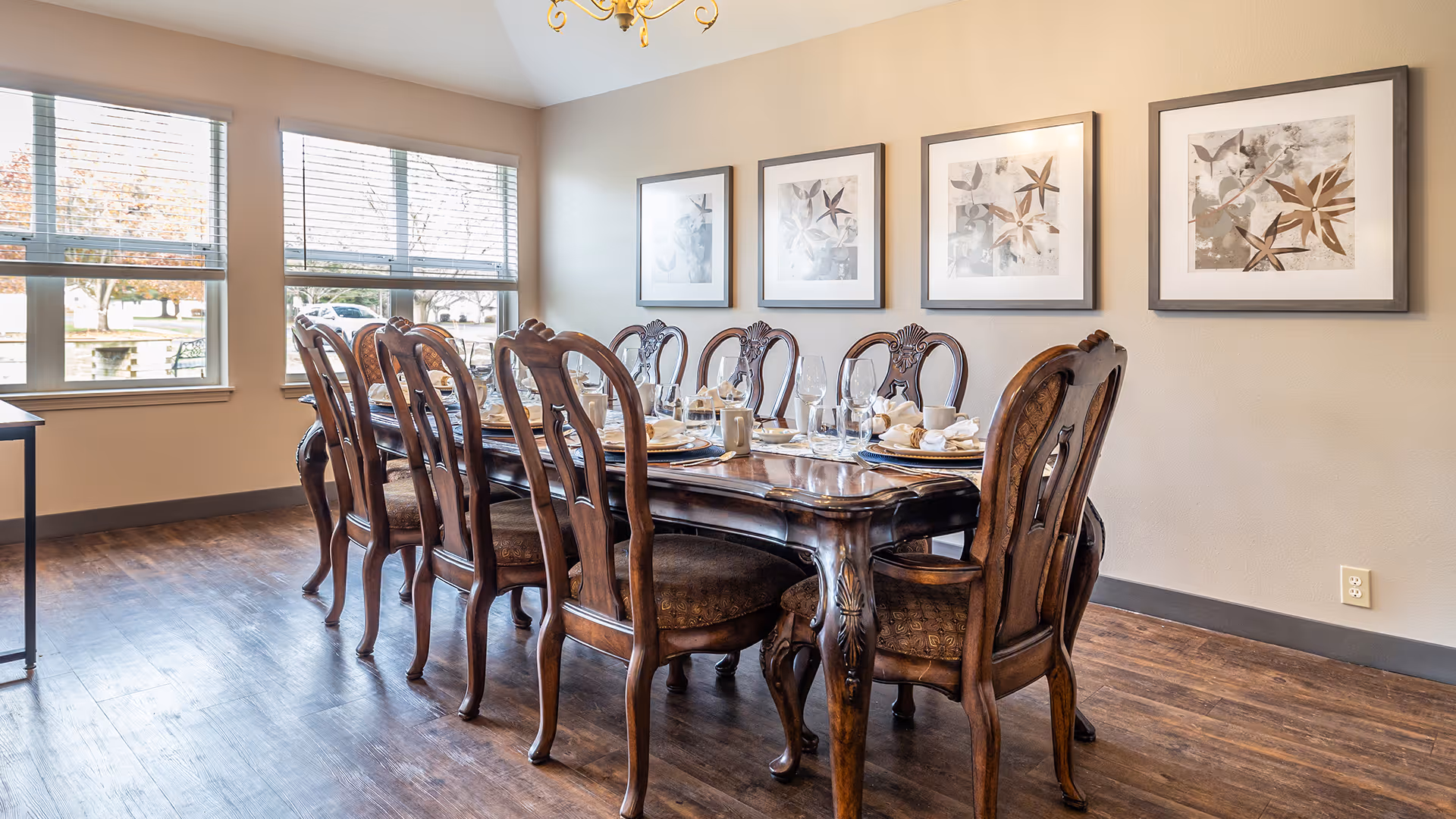 A dining room with a long wooden table set for eight people with plates, glasses, and napkins. The room has large windows with blinds, wooden flooring, a chandelier, and four framed botanical prints on the wall.