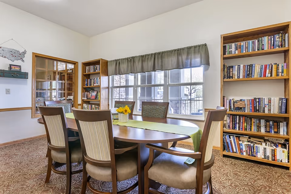 A cozy room with a wooden oval table surrounded by six cushioned chairs. The table has a green runner and a small vase with yellow flowers. The room features two wooden bookshelves filled with books, a large window with a green valance, and a carpeted floor with a patterned design. A decorative map and a small shelf are mounted on the wall.